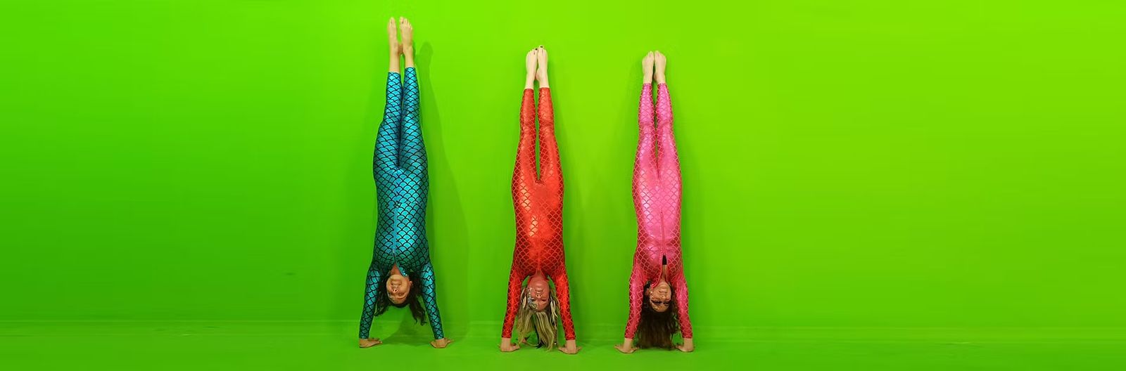 Three aerialists in sparkling costumes doing handstands against a green wall