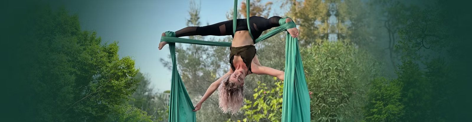 Aerialist hanging upside down in a split between green silks among the trees