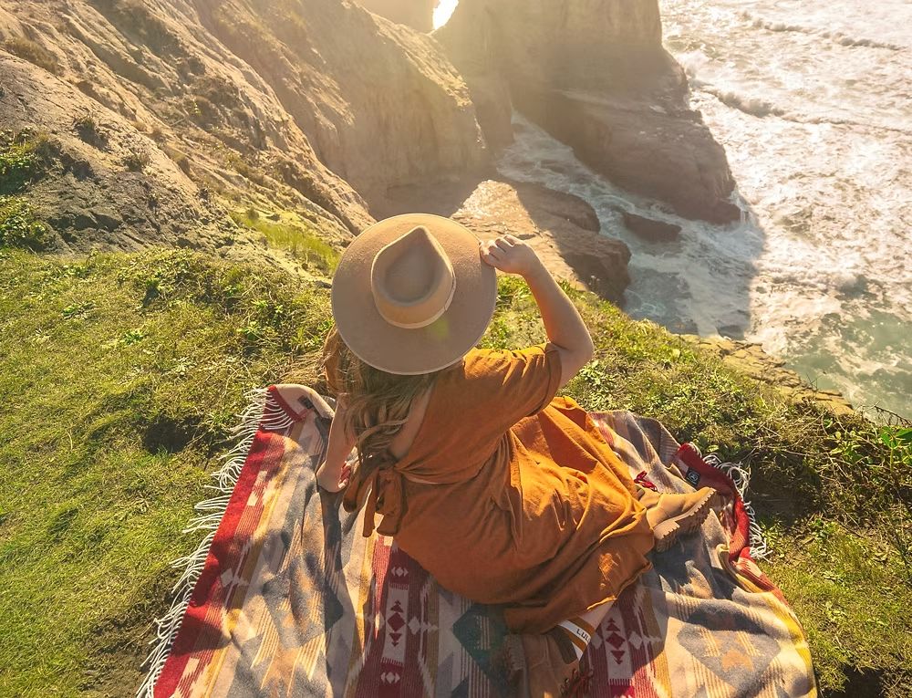 A hiker in a wide-brimmed hat resting on a blanket above the sea cliffs